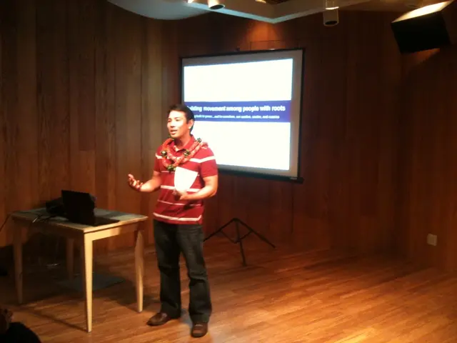 He is standing on a stage. He's holding a paper. His talking. There is a table. There is a...