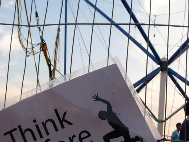 In this picture we can see a paper on the fence, in the background we can find few people and metal...